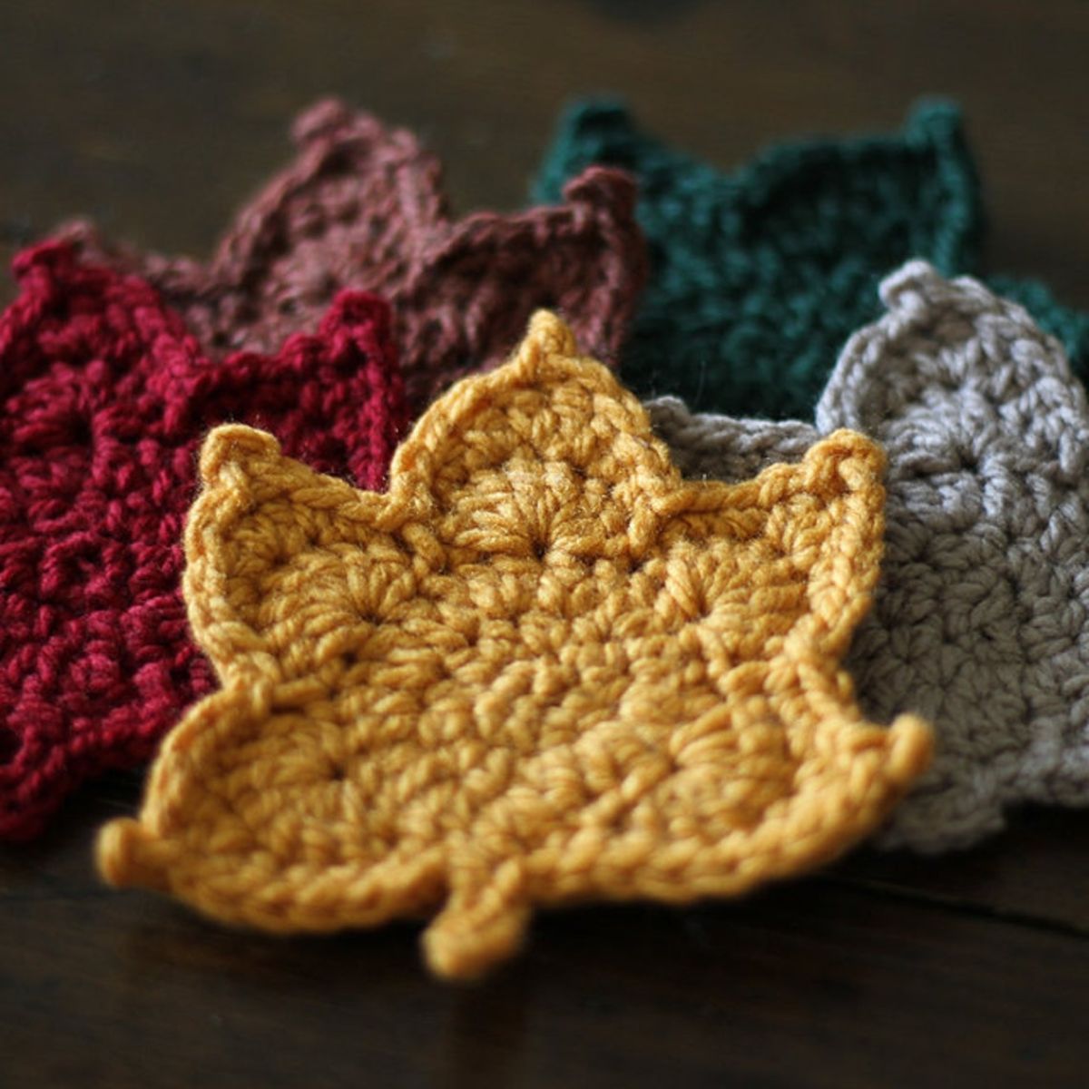 A small pile of yellow, gray, red, brown, and green crochet maple leaves lying on a dark brown background.