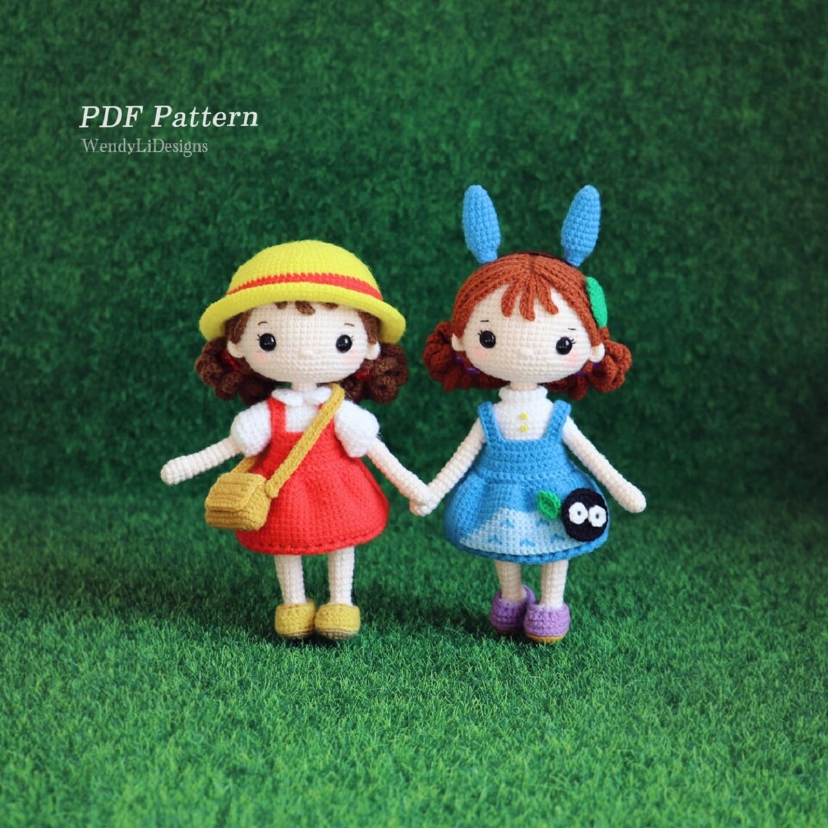 Crochet stuffed Mei and Satsuki wearing a red and blue dress holding hands on a green artificial grass background.