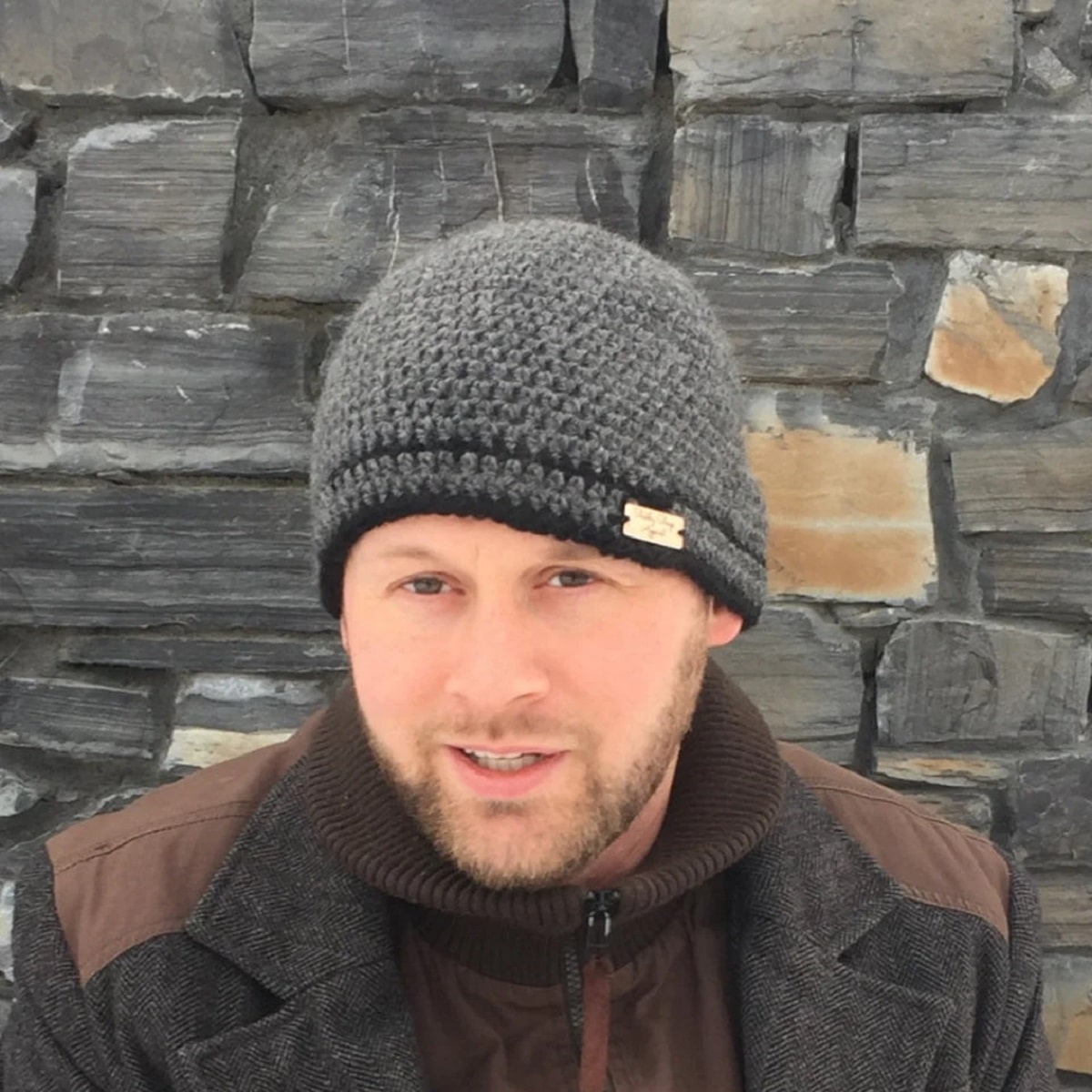  Man with a goatee standing in front of a stone wall wearing a gray beanie hat with a black band around the bottom.