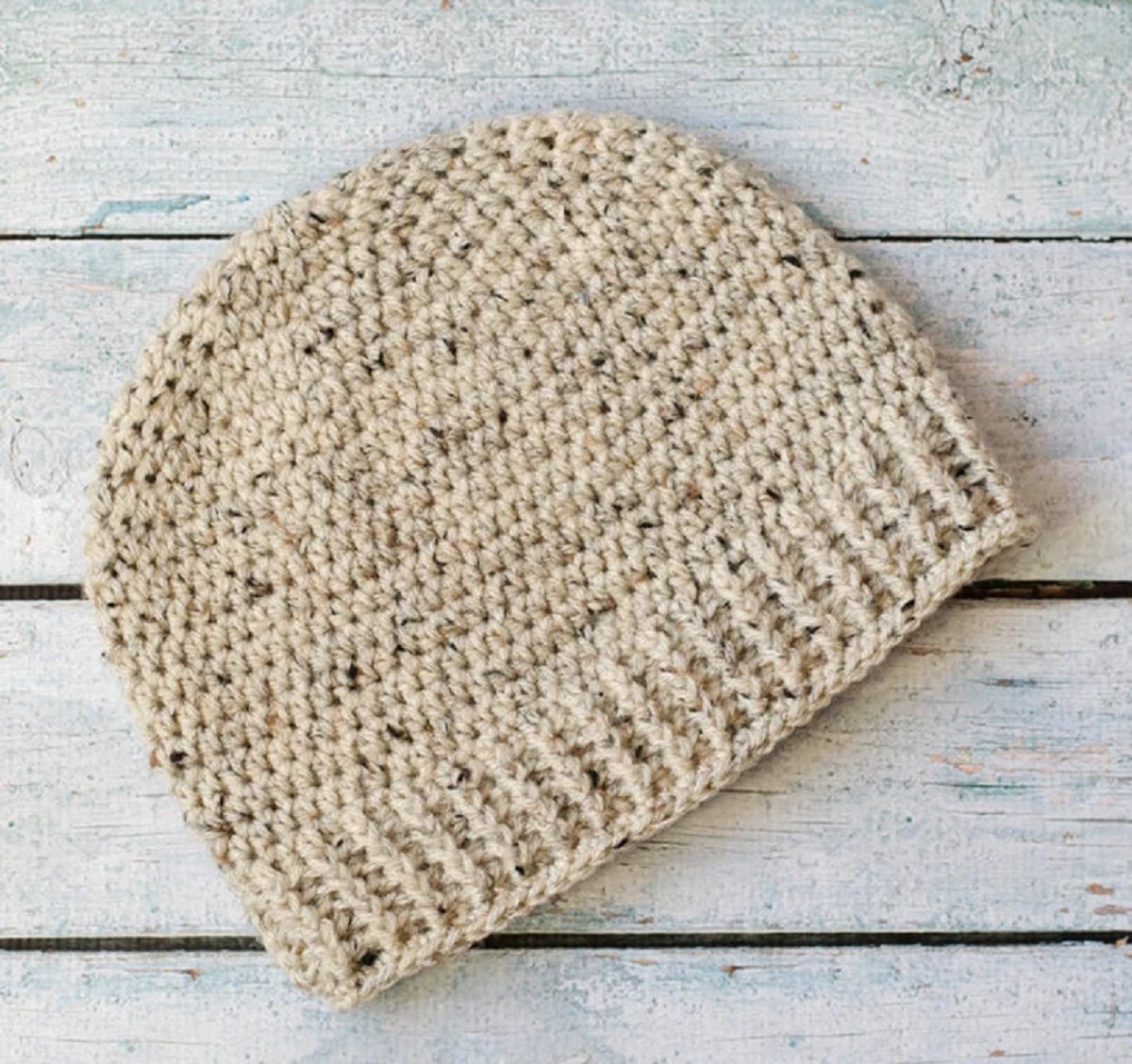 A cream crochet beanie hat with blue and green speckles throughout and a thick band on the bottom on a white wooden floor.