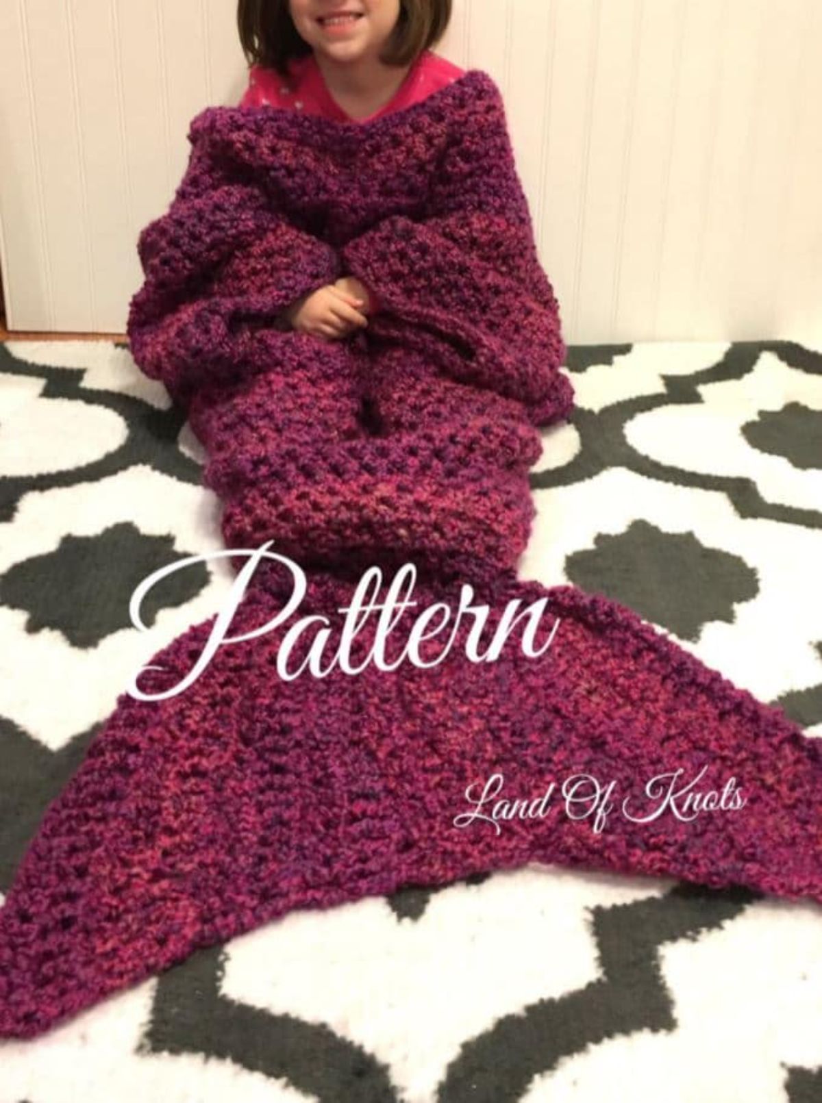 Dark purple crochet blanket with mermaid tail worn by a smiling brunette woman sitting on a black and white carpet. 