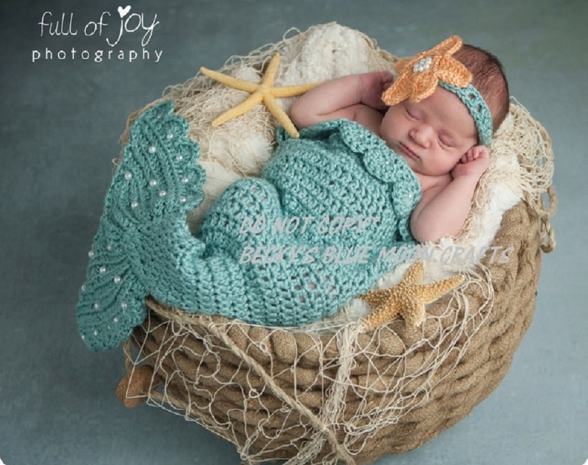  Newborn baby sleeping in a wicker basket with a cream blanket wearing a green crochet mermaid costume and matching headband.