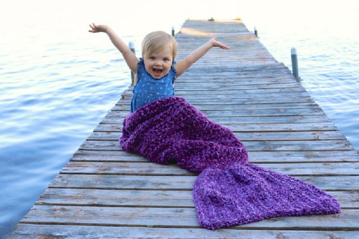 A Blonde child sitting on a pier wearing a blue top and oversized purple crochet mermaids tail blanket.