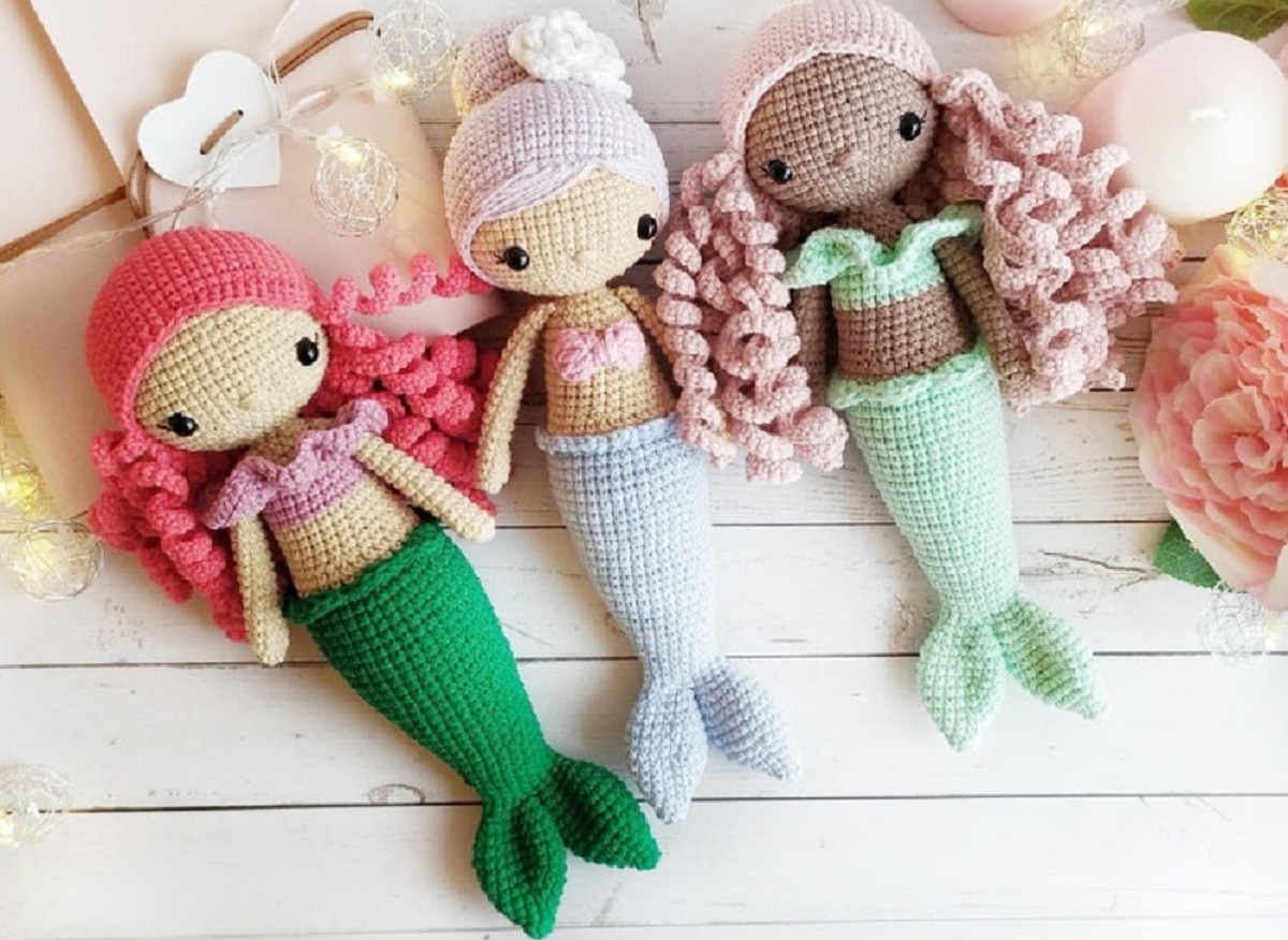 Three crochet mermaid dolls in a line with green and blue tails with pink and purple hair on a white wooden background.