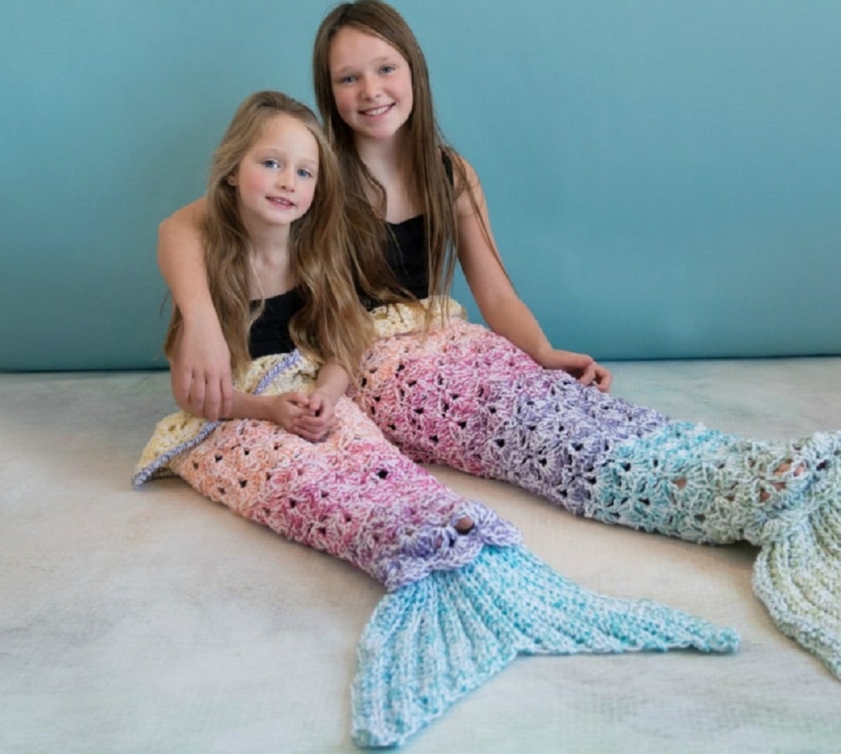  Two young girls sitting next to each other wearing multicolored pastel crochet mermaid tail blankets from their waists to their feet.