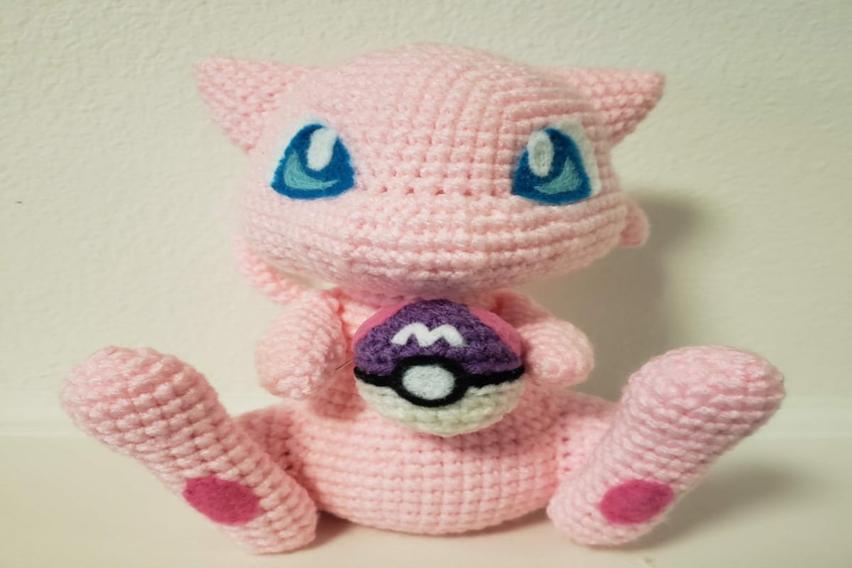 Pale pink Mew sitting on a white background with blue eyes, dark pink circles on its feet and holding a purple and white Pokeball in its hands.