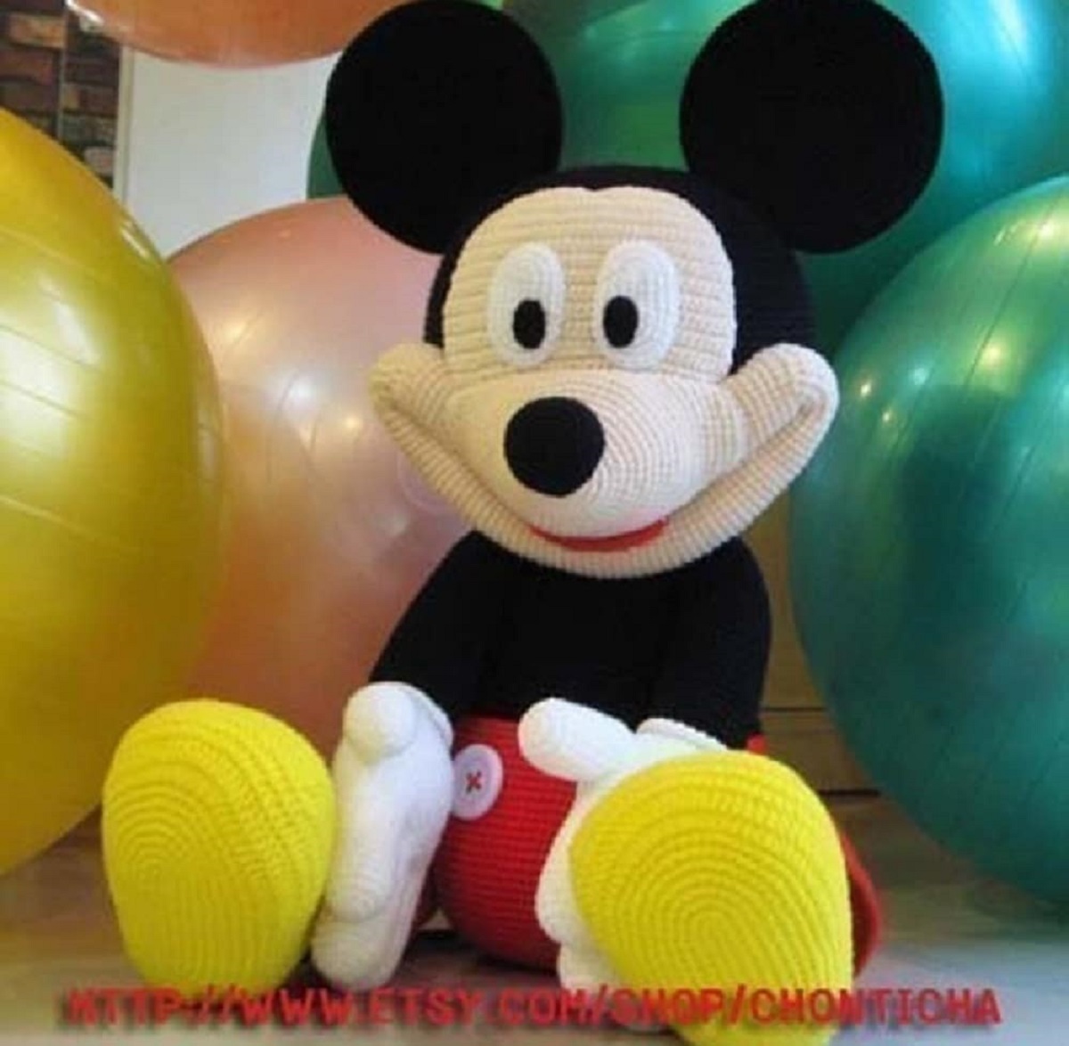 Stuffed crochet Mickey Mouse toy with a large smile and Mickey’s traditional outfit sitting next to balloons. 
