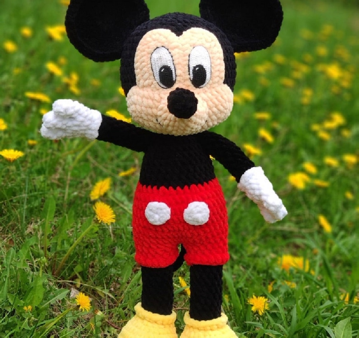 Stuffed crochet Mickey Mouse wearing red and white shorts and yellow shoes standing with one arm raised on some grass.