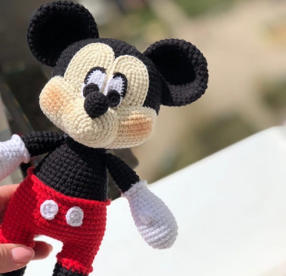 Crochet stuffed Mickey Mouse toy wearing red shorts with two shite circles being held by one hand.