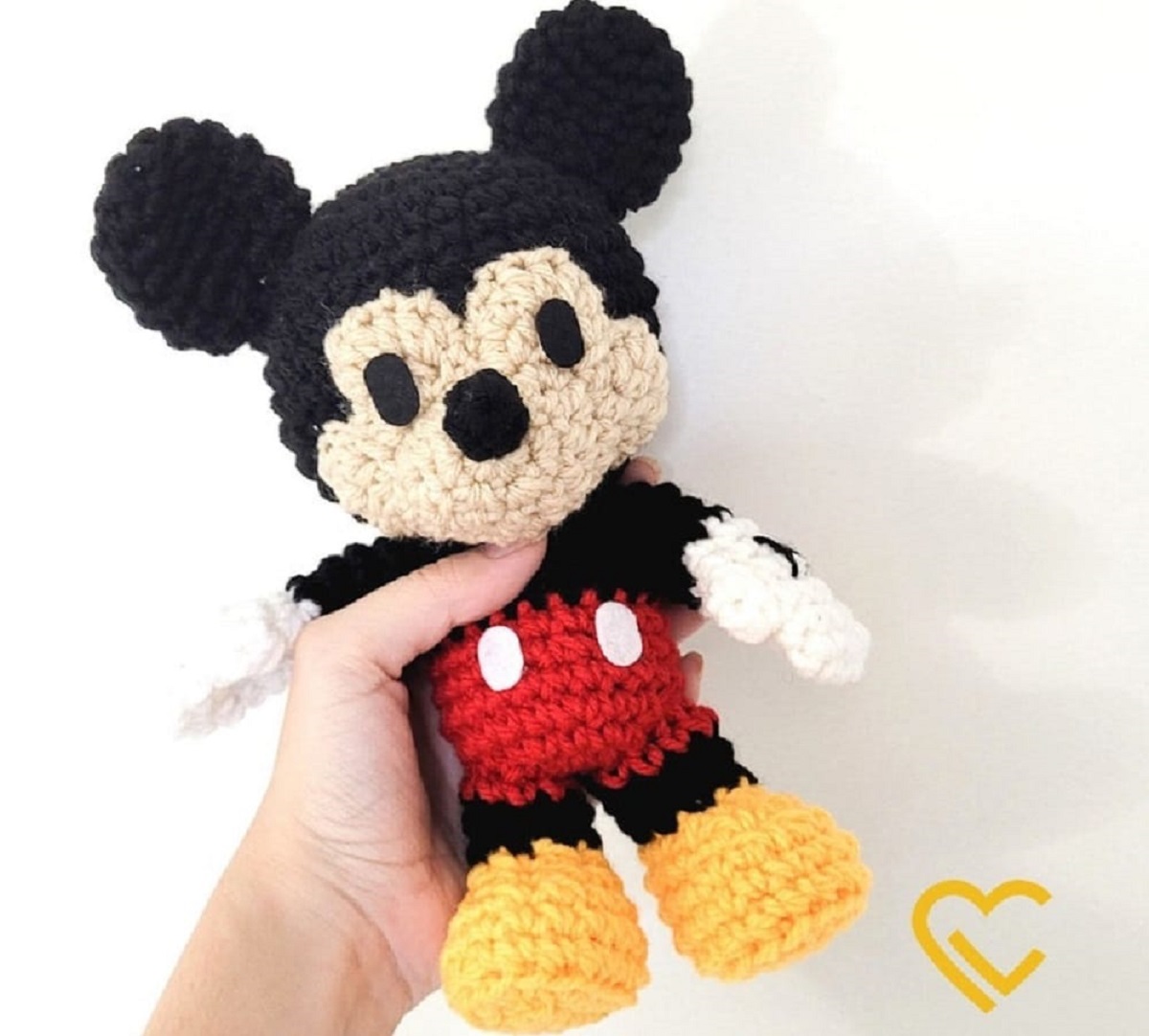 Mini crochet Mickey Mouse toy with black eyes and a nose wearing red shorts with white spots and yellow shoes helped by a hand.