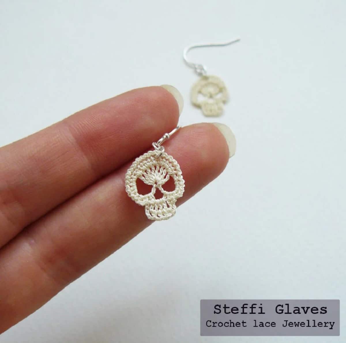 Two fingers with a small crochet skull earring on them. The other earring can be seen underneath on the white background.