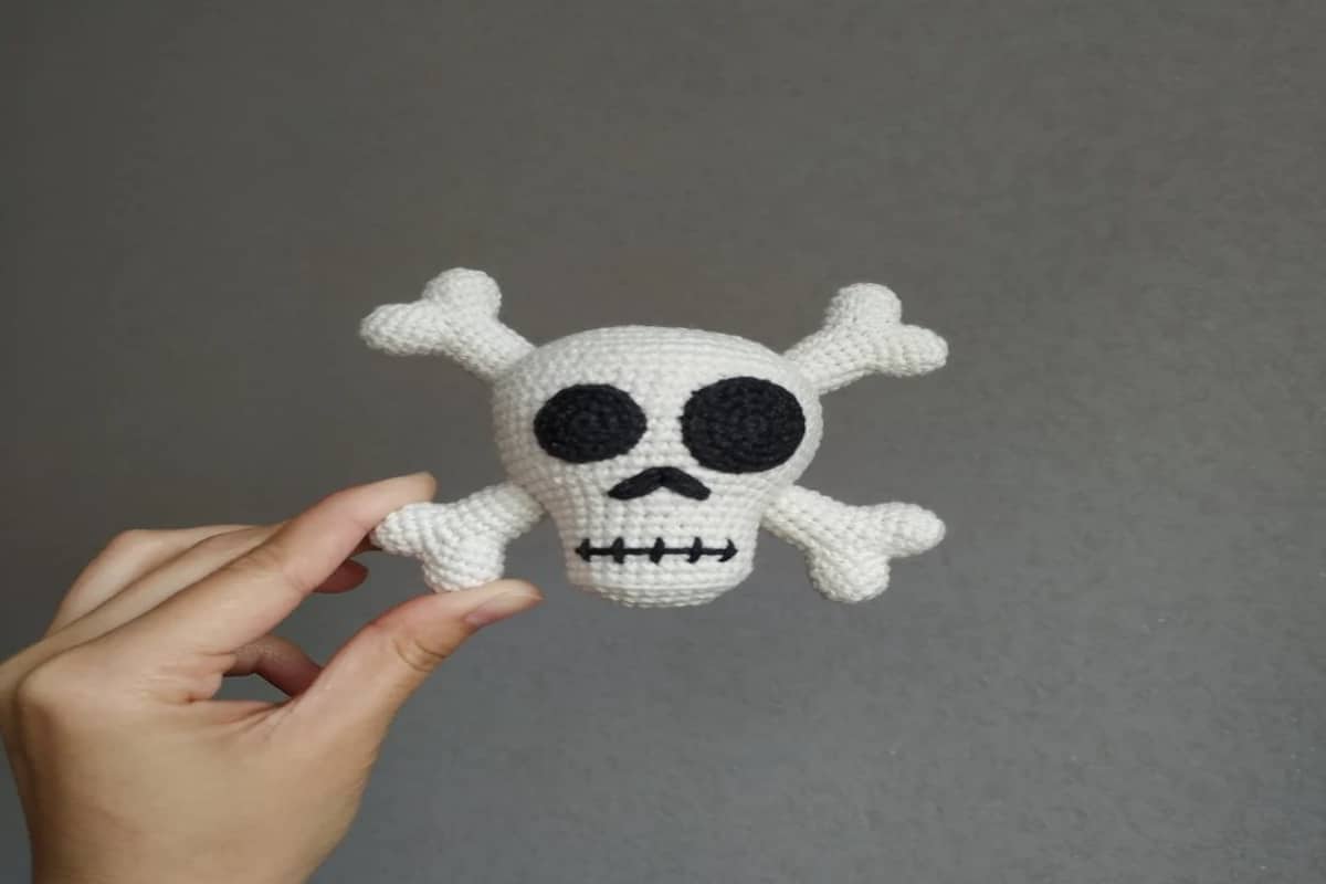 Small white crochet skull and bones with black eyes, nose, and mouth held up by a hand.