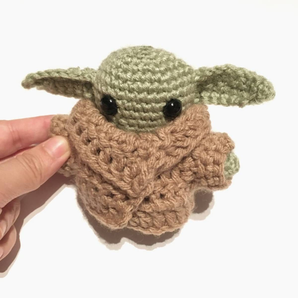 Small crochet baby Yoda in a brown robe held by a hand on a white background.