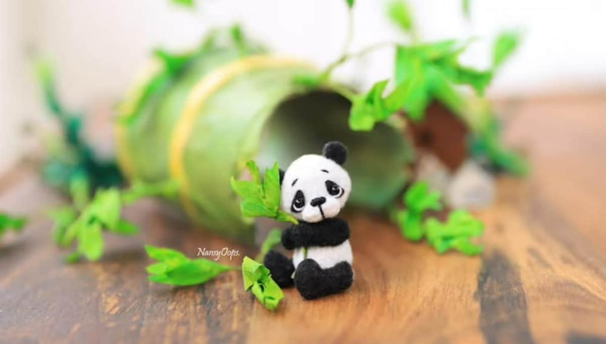 Mini crochet black and white panda sitting down holding some green leaves sitting in front of other leaves and branches. 