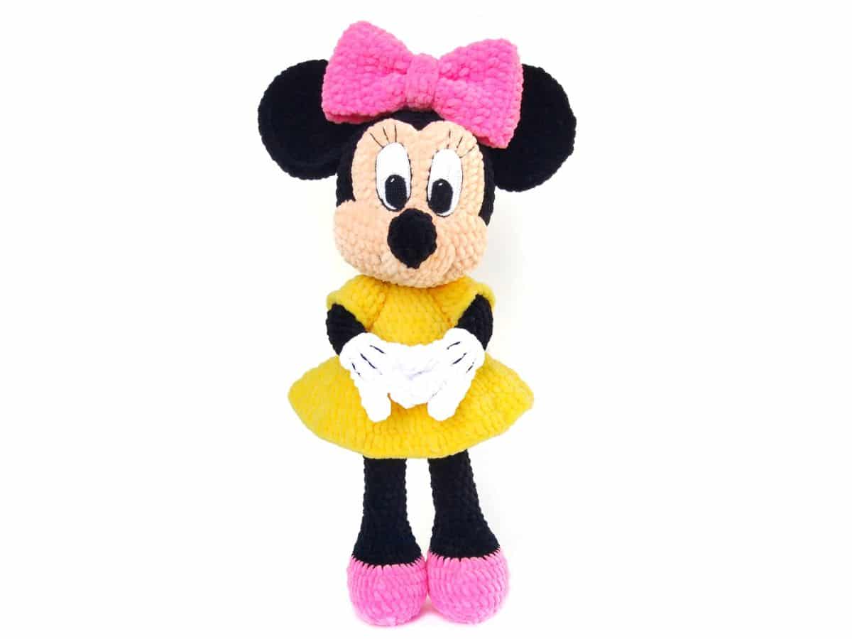 Small crochet Minnie Mouse toy in a yellow dress with a pink bow on her head and pink shoes standing on a white background