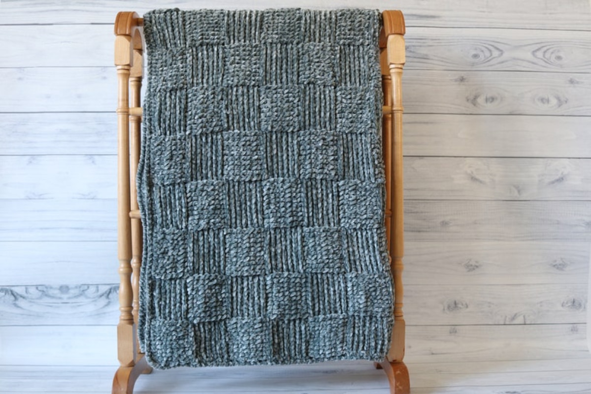 Green crochet throw using a large basketweave pattern draped over a wooden hanger on a pale wooden background.