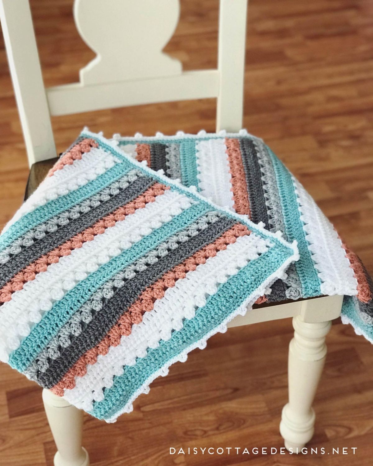 A small crochet blanket with blue, white, orange, navy, and pale gray horizontal stripes with a small white trim on all sides, placed on a cream chair.
