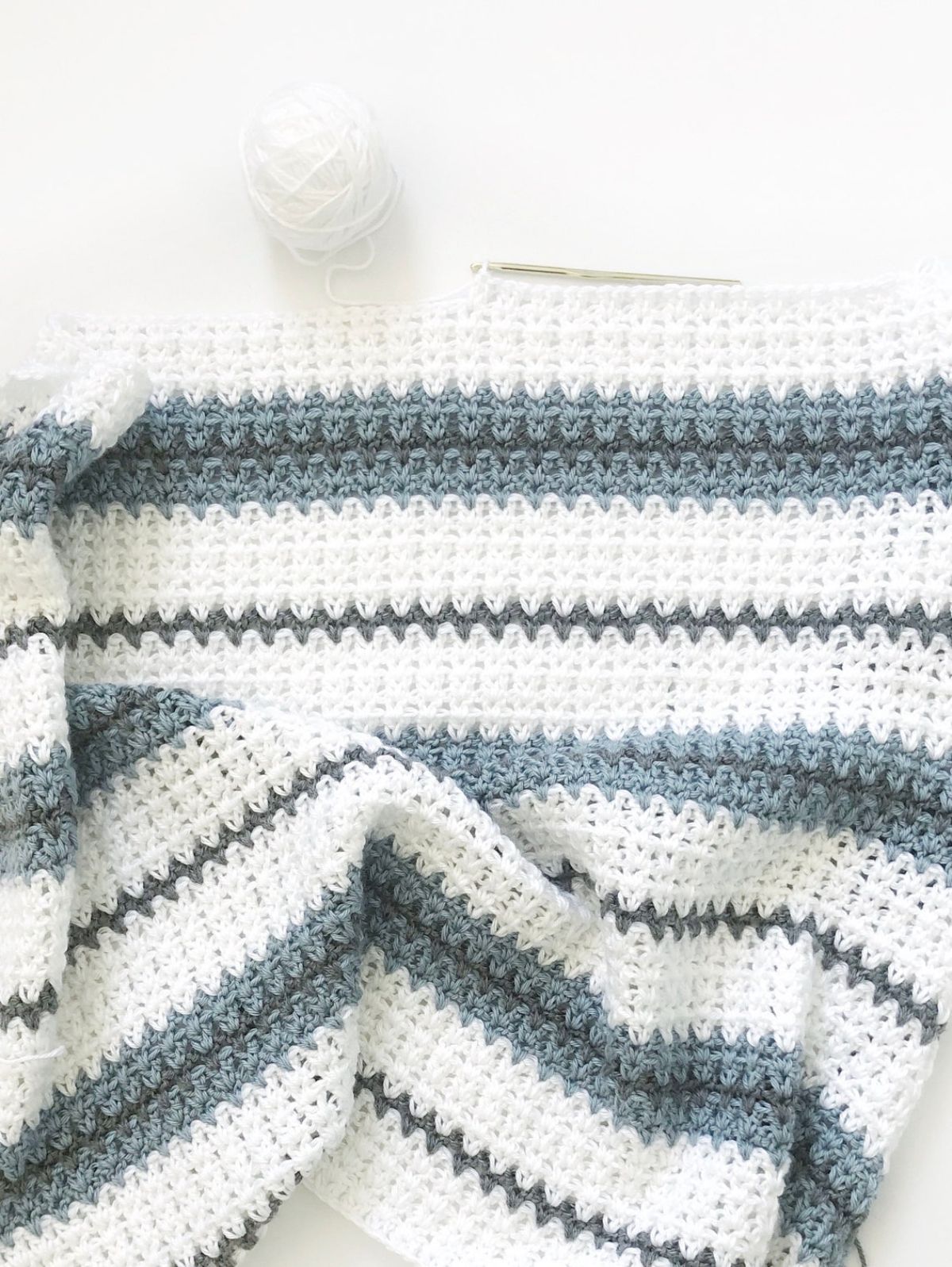 A white crochet blanket with a thin dark green stripe across it and thicker blue stripes next to some white yarn.