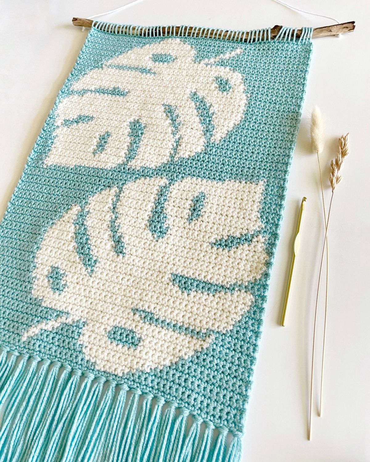 An aqua colored crochet hanging piece with large white leaves and blue tassels hung on a thin stick with a crochet hook and white leaves next to it.