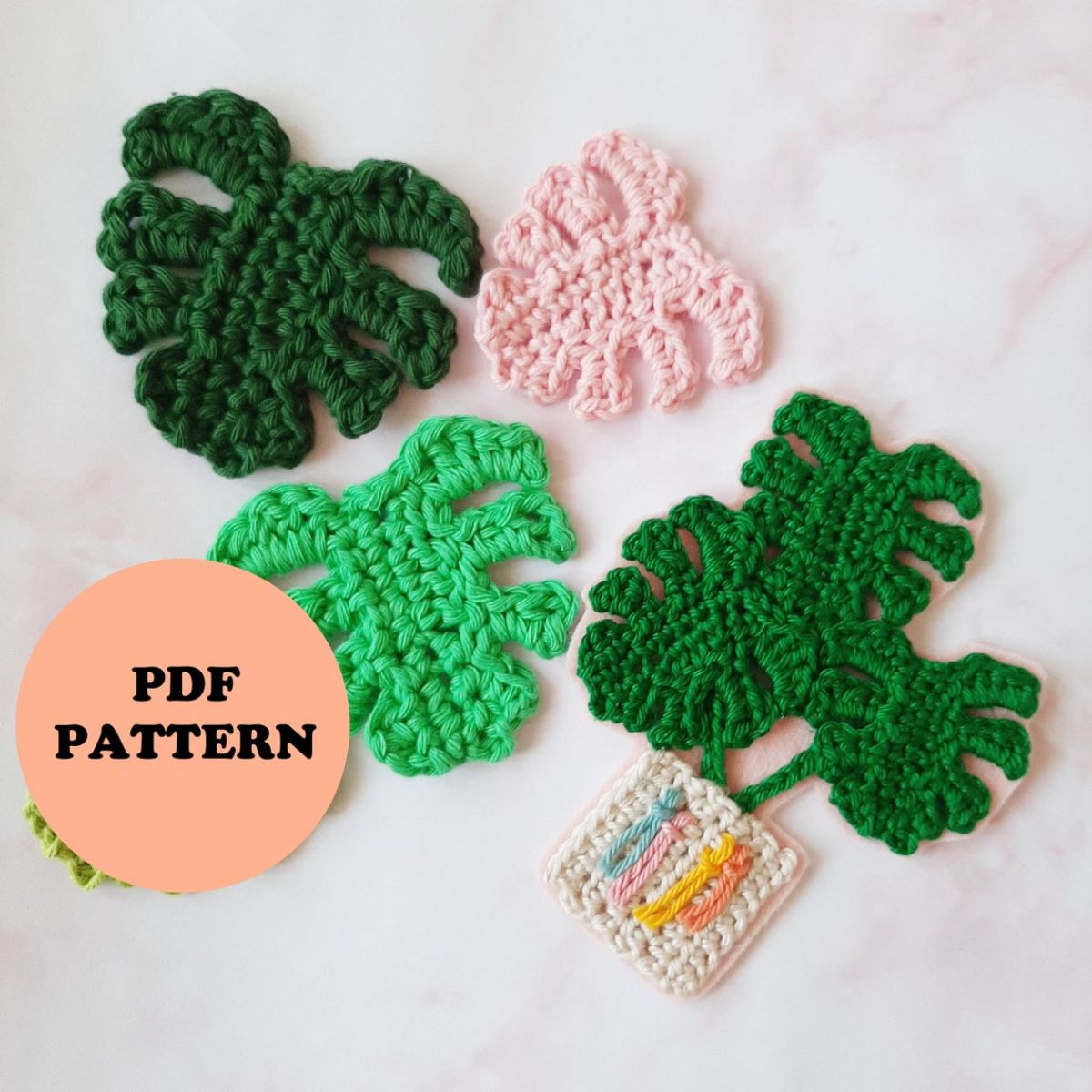 Four crochet monstera leaves in dark green, green, and pink scattered on a light background with one leaf coming out of a white pot.