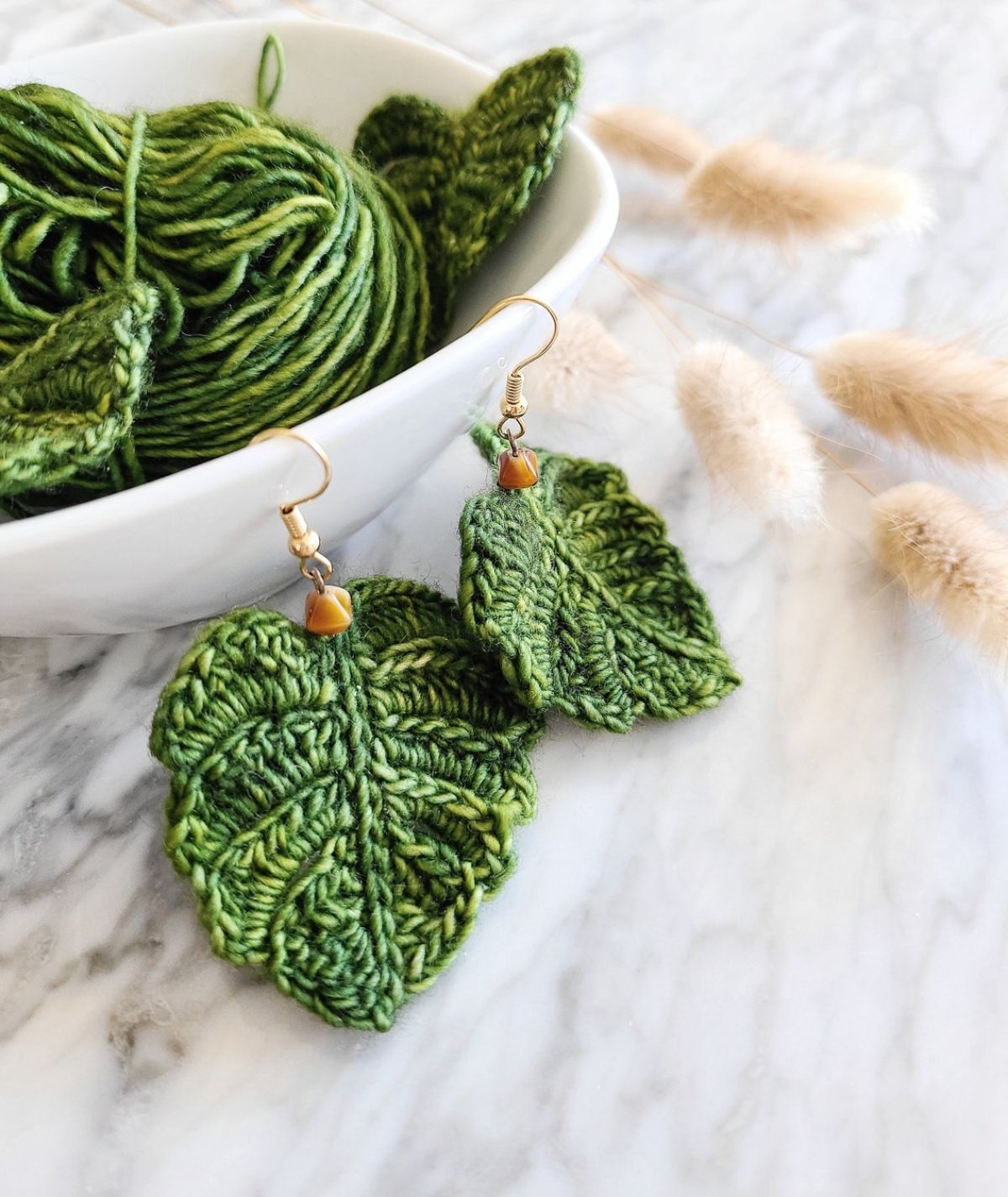 A small pair of green crochet monstera leaf earrings hanging from a white bowl filled with the same color yarn.