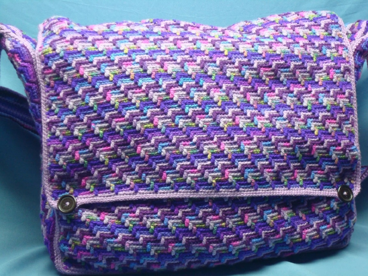 A satchel style bag with a purple, pink, yellow, blue, and green trail of tears pattern across it and two buttons at the front to secure the bag.