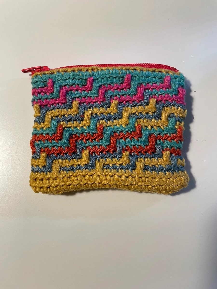 A pink, yellow, red, blue, and green crochet purse with a mosaic pattern and an orange zipper at the top.