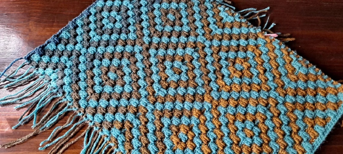 An orange, brown, and blue diamond pattern crochet rug with tassels along the bottom on a dark wooden floor.