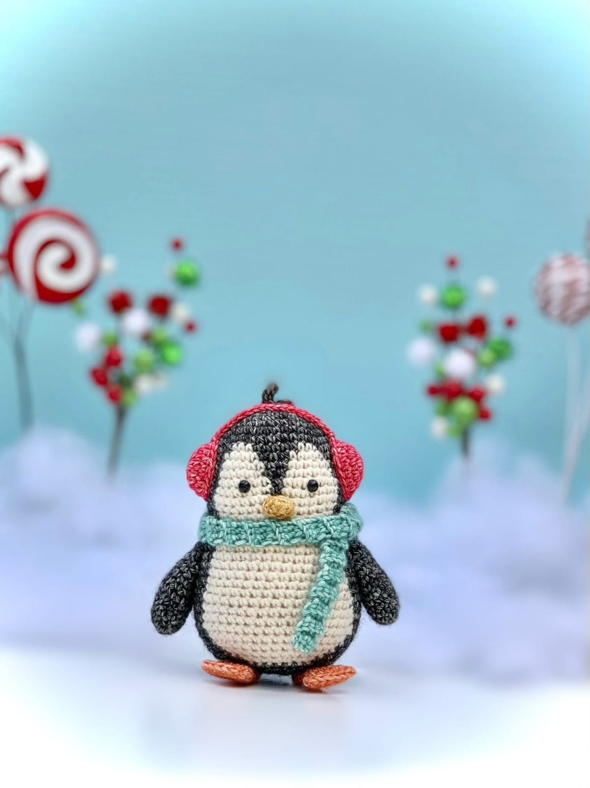 Small crochet black and white penguin wearing a blue scarf around its neck and red earmuffs in front of a snowy background with candy canes.