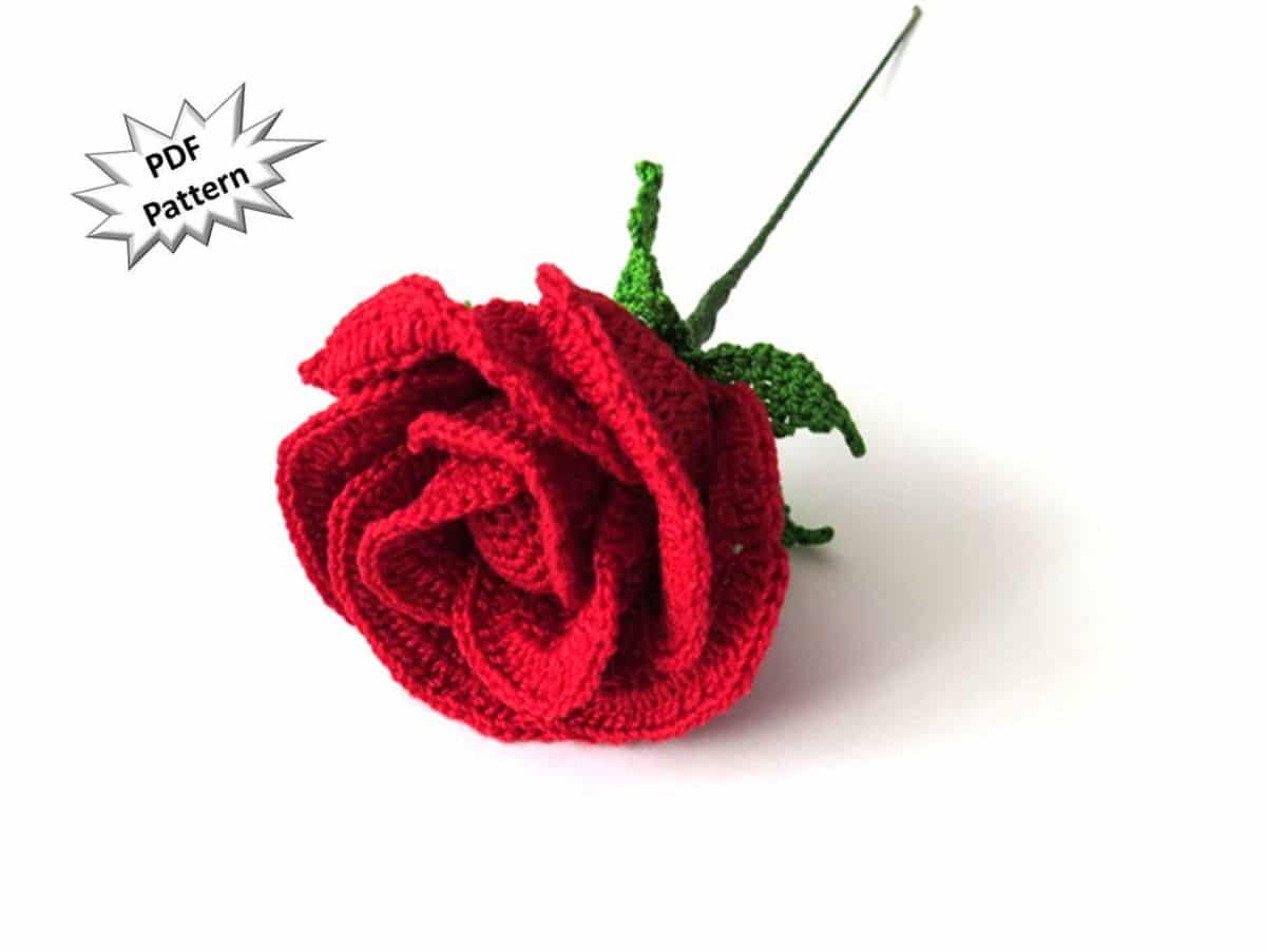 A red crochet rose laying on a white background with two green leaves underneath and a thin stem attached.