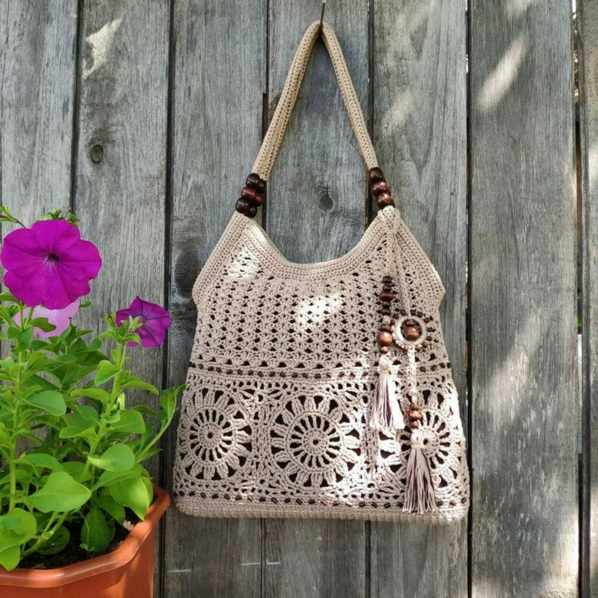 A beige crochet bag with a large flower print running along the bottom, some beads dangling from the right side, and a thick strap at the top.