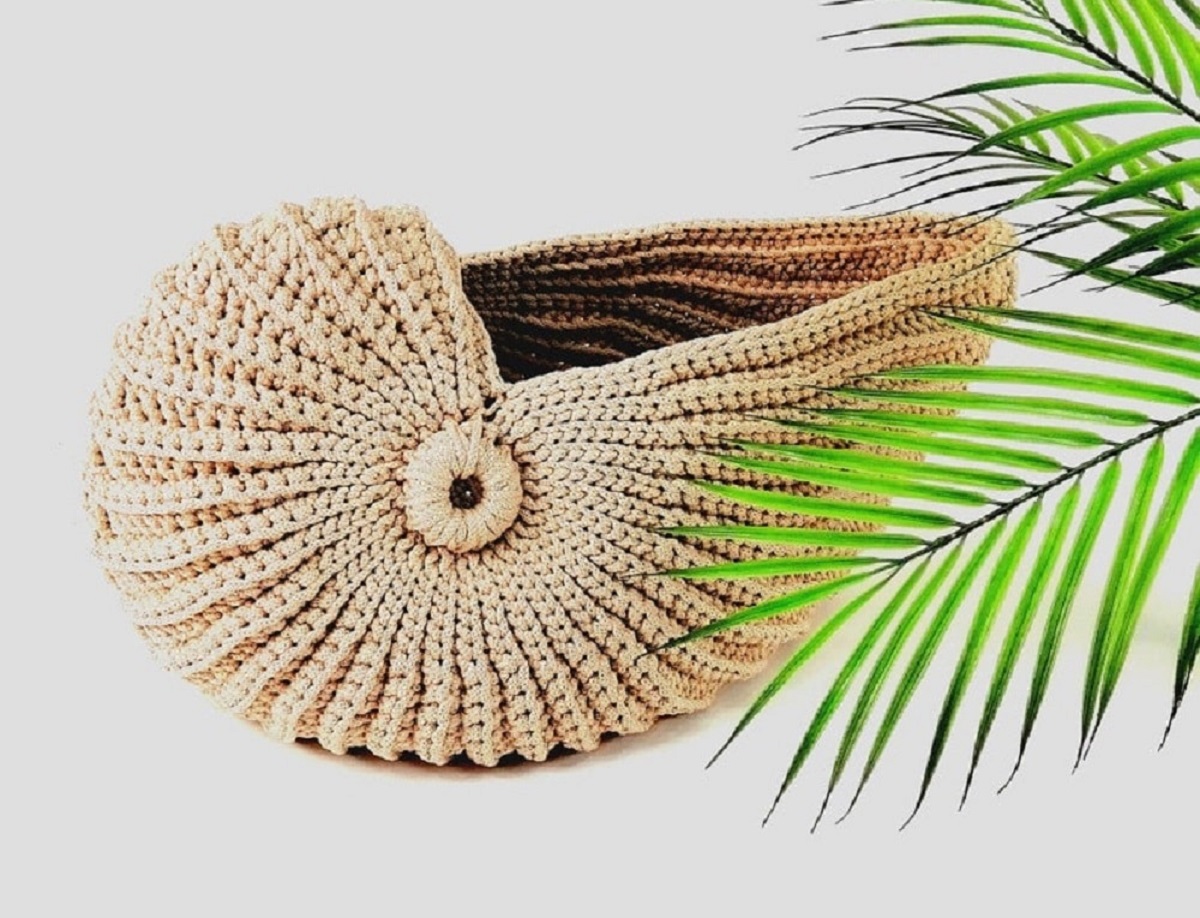 A beige colored crochet shell with the top open to store items in on a white background next to green leaves.