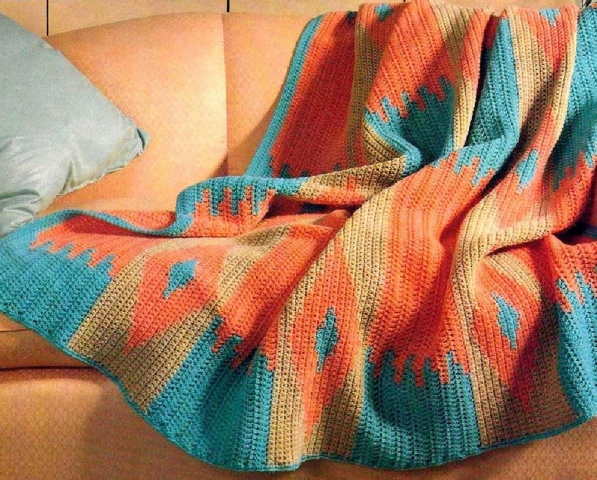 Blue, orange, and yellow crochet blanket with beautiful sunset colors draped over an orange sofa. 