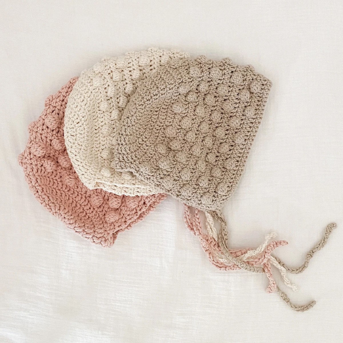 Beige, white, and pale pink crochet bonnets lying on top of each other with small circles stitched in and braided string to secure the bonnets.
