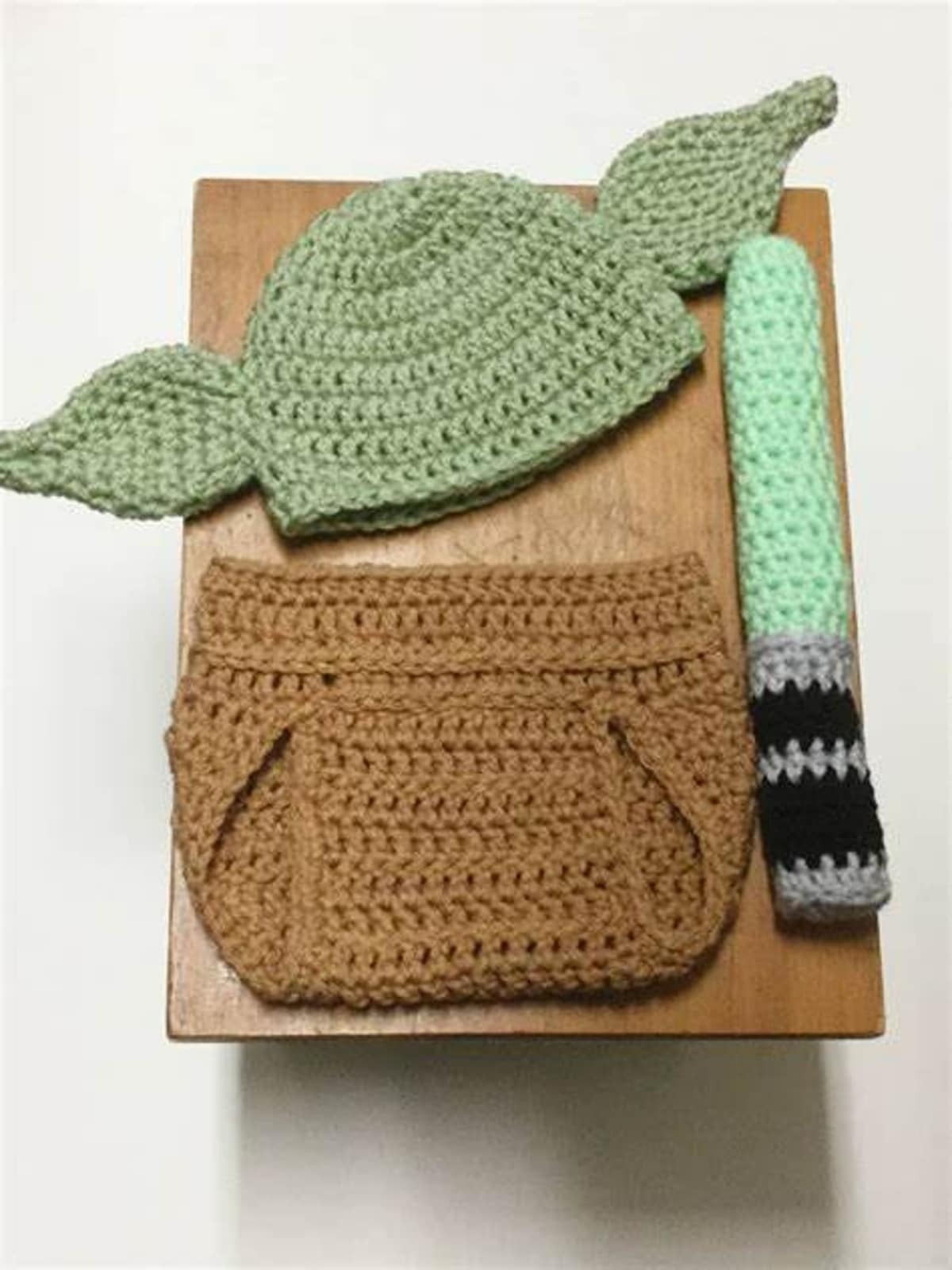 Newborn crochet baby Yoda costume with a green hat, brown diaper cover, and green tail with gray and black stripes on the bottom.