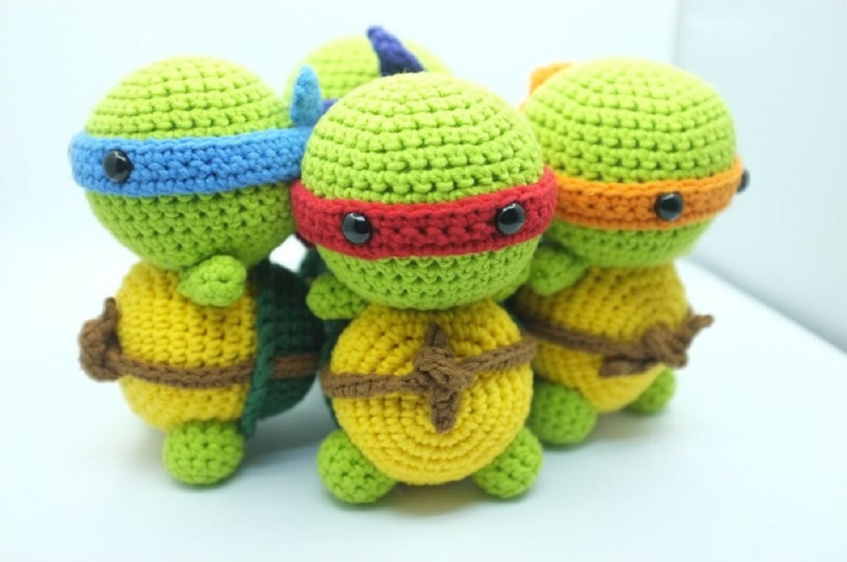 Four crochet ninja turtles standing in a circle with green bodies, yellow bellies, and red, orange, and blue eye masks on.