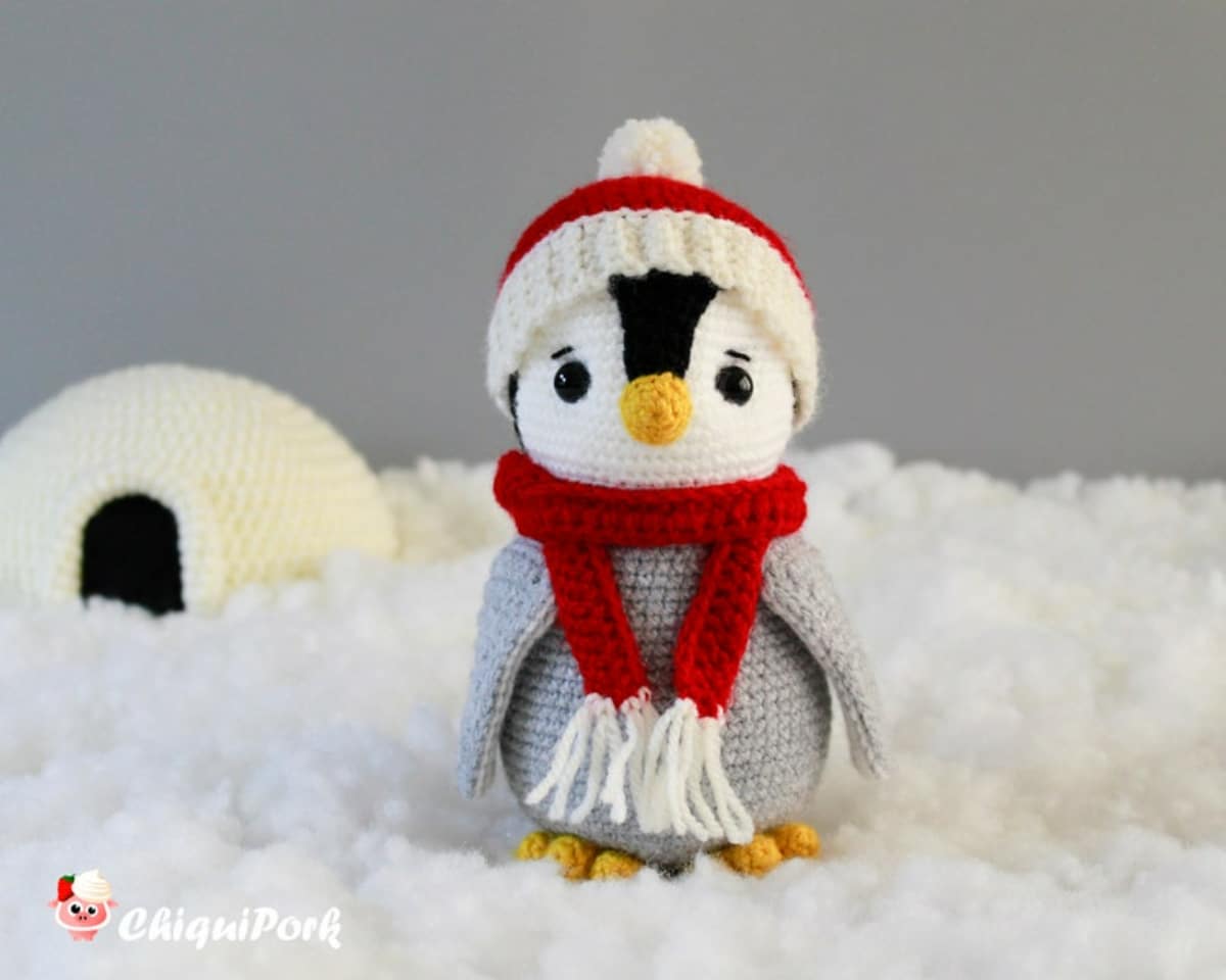 White crochet penguin wearing a red and white bobble hat, red and white scarf, and a grey jacket standing on some fake snow by an igloo.