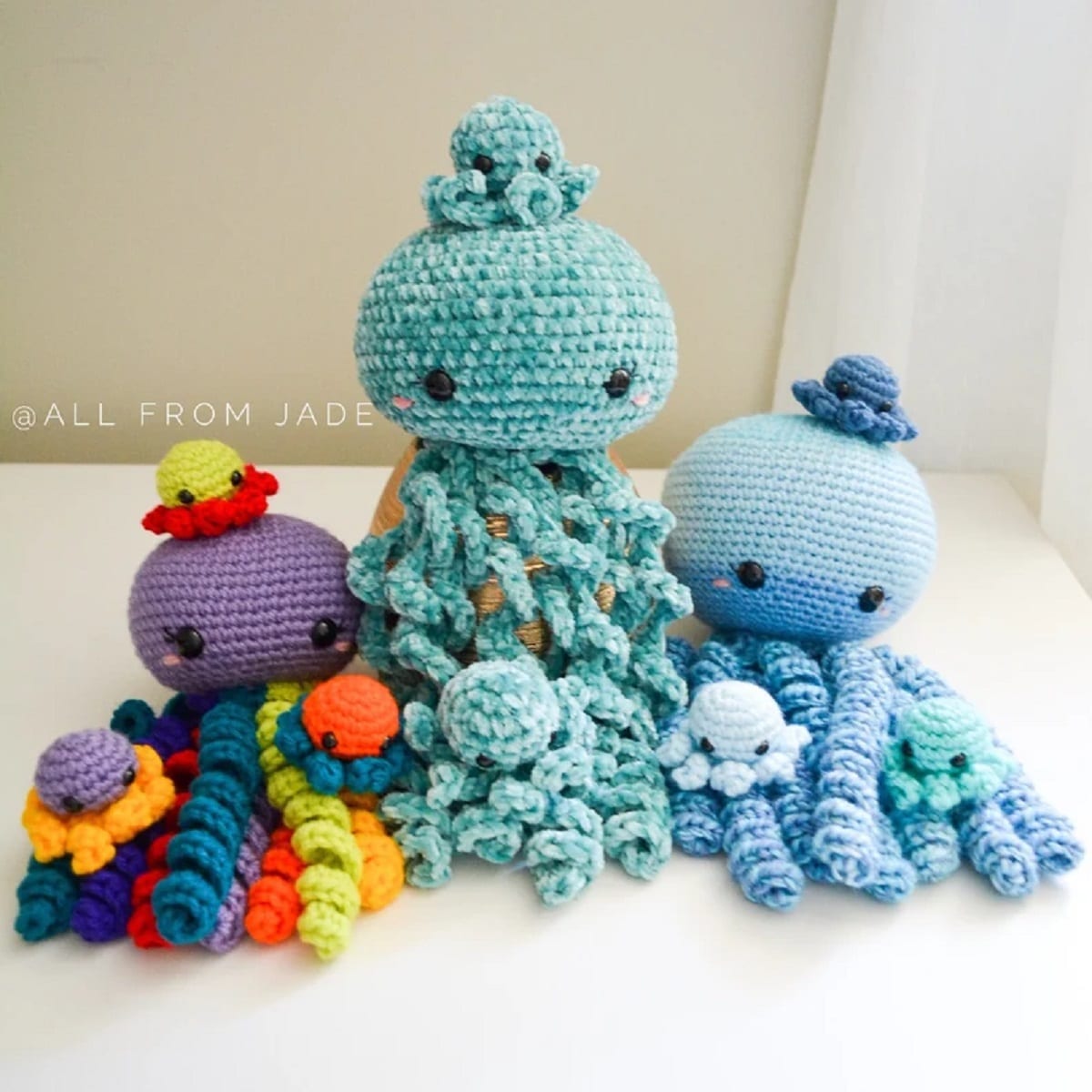 Large textured crochet octopuses in blue and purple with ruffle style tentacles sitting on a white floor with mini octopus babies around them.