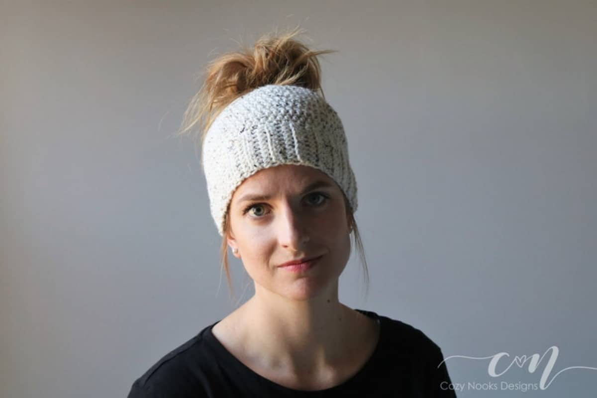 A blond woman wearing a white crochet hat with a large blond messy bun pulled through the top.