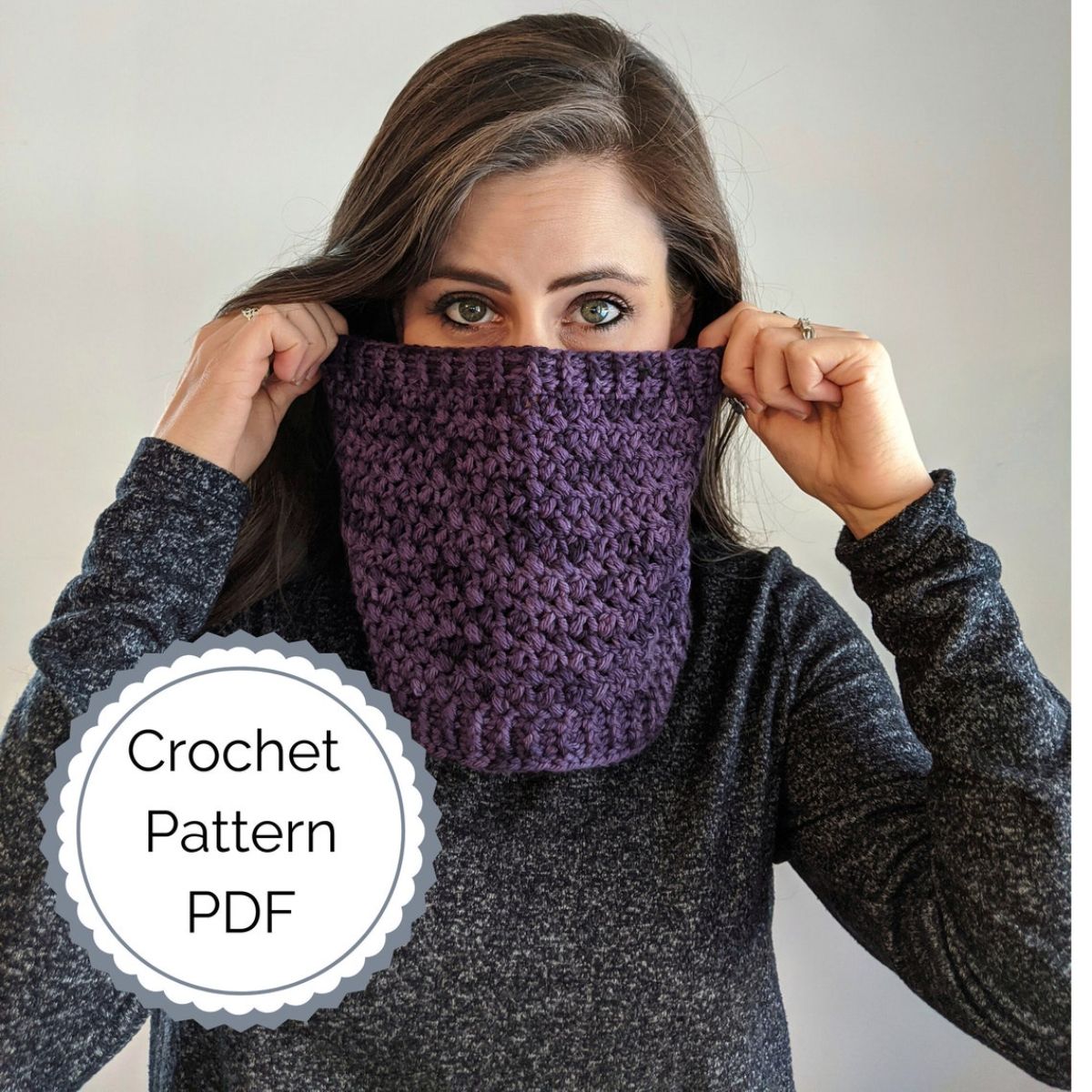 Brunette woman with a purple crochet cowl over her neck with her hands holding it to cover her nose and mouth too.