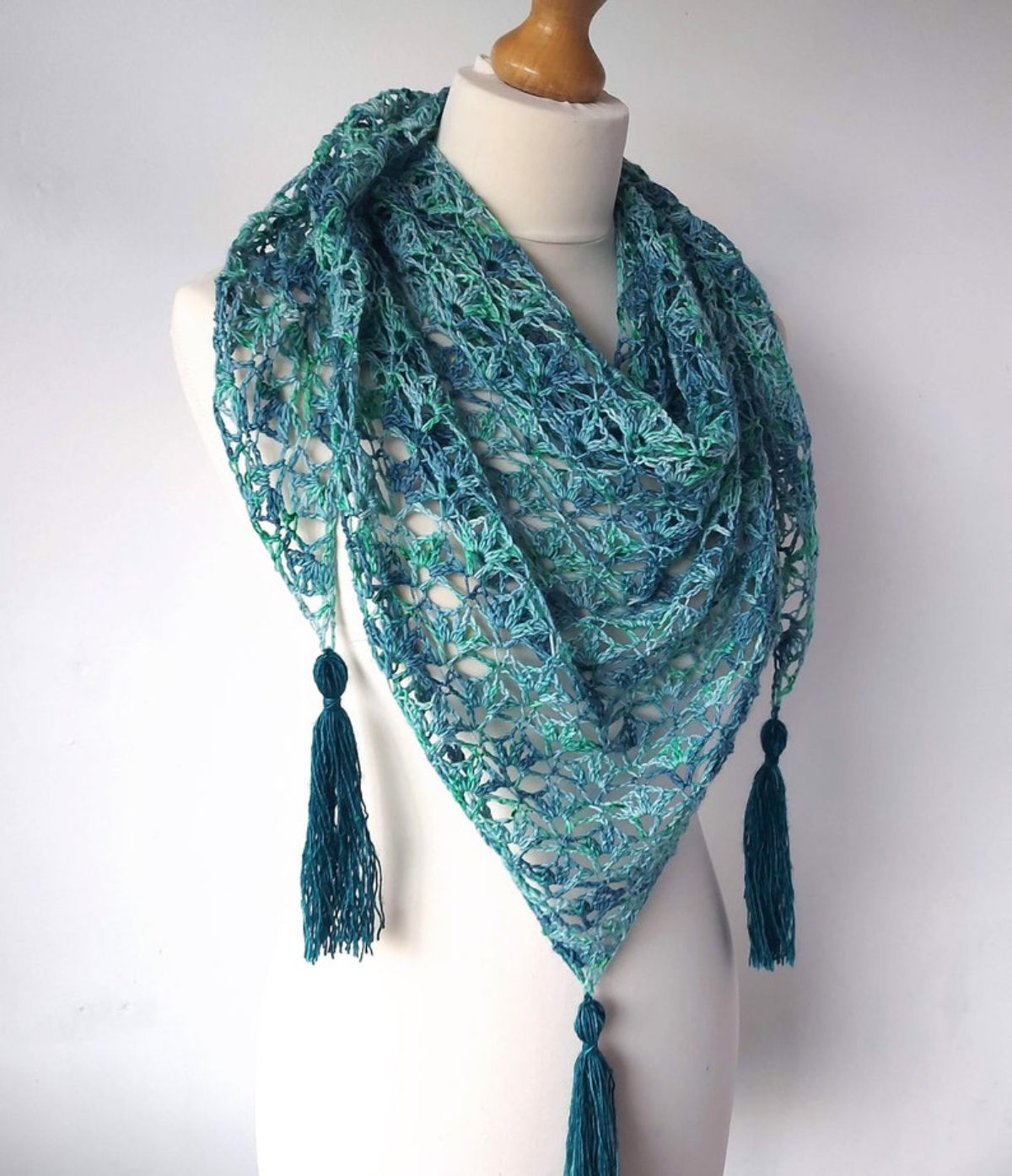 Light and dark green lace style crochet skein shawl wrapped around a mannequin with three large dark green tassels hanging off each corner. 