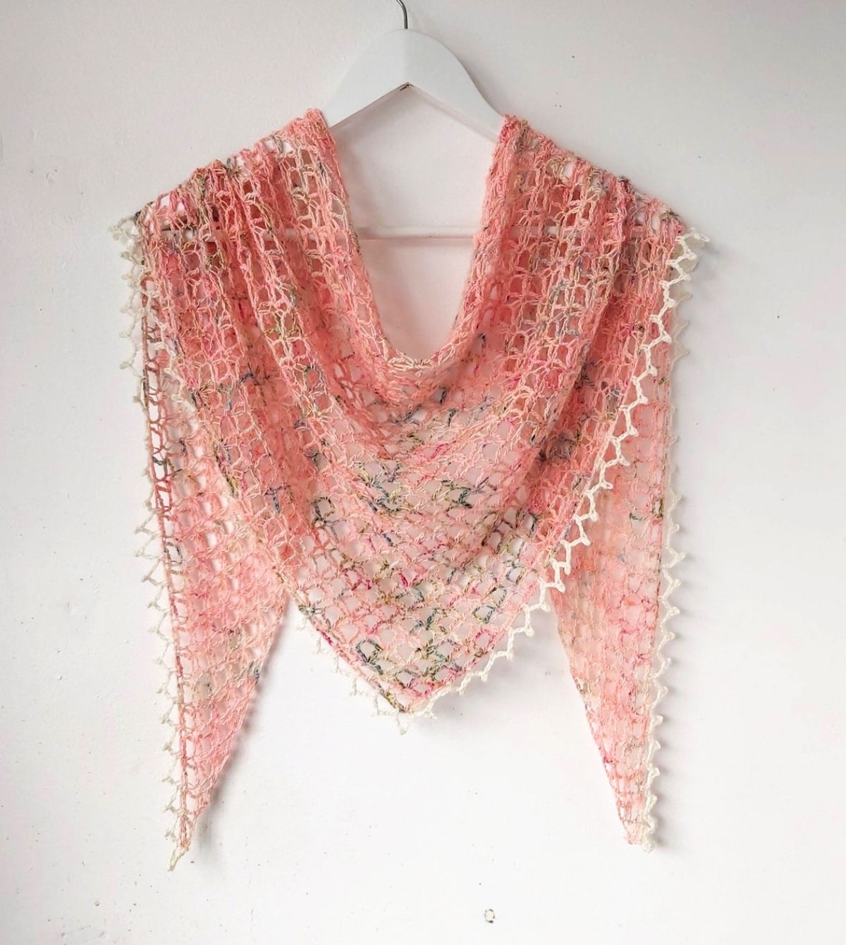 Pink and cream ombre style crochet skein diamond shaped shawl with a lace style trim hanging over a cream wooden hanger. 