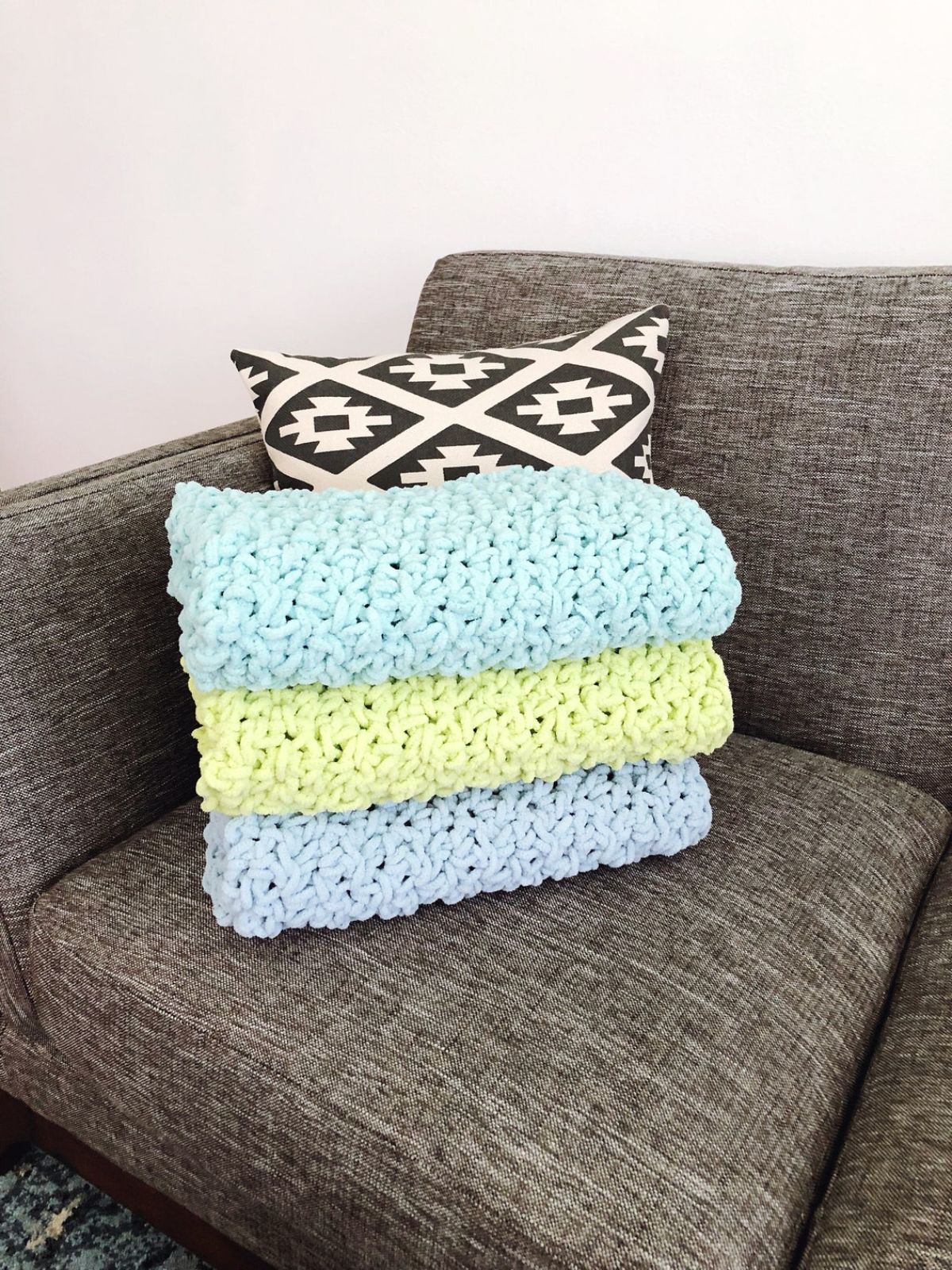 Three skein style crochet blankets folded on top of each other on a grey sofa. The blankets are gray, yellow, and light blue.