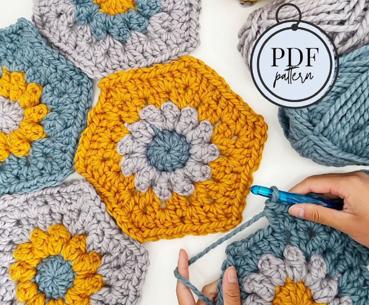 Large yellow, blue, and gray crochet hexagons with a daisy in the center next to some hands and a crochet hook finishing one of these hexagons.