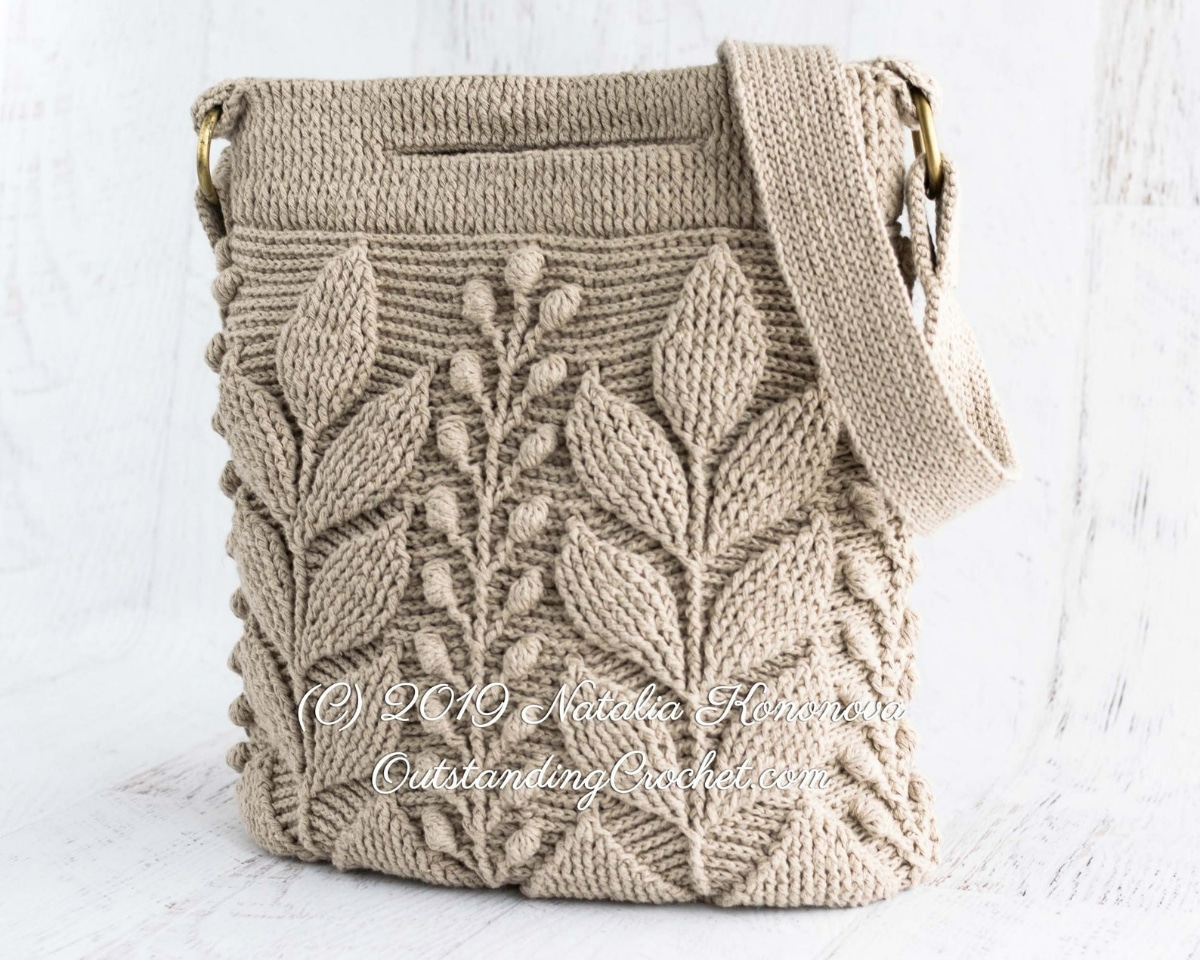 Beige crochet bag with leaves stitched from the top to bottom with a thick band across the top and a strap draped over it.