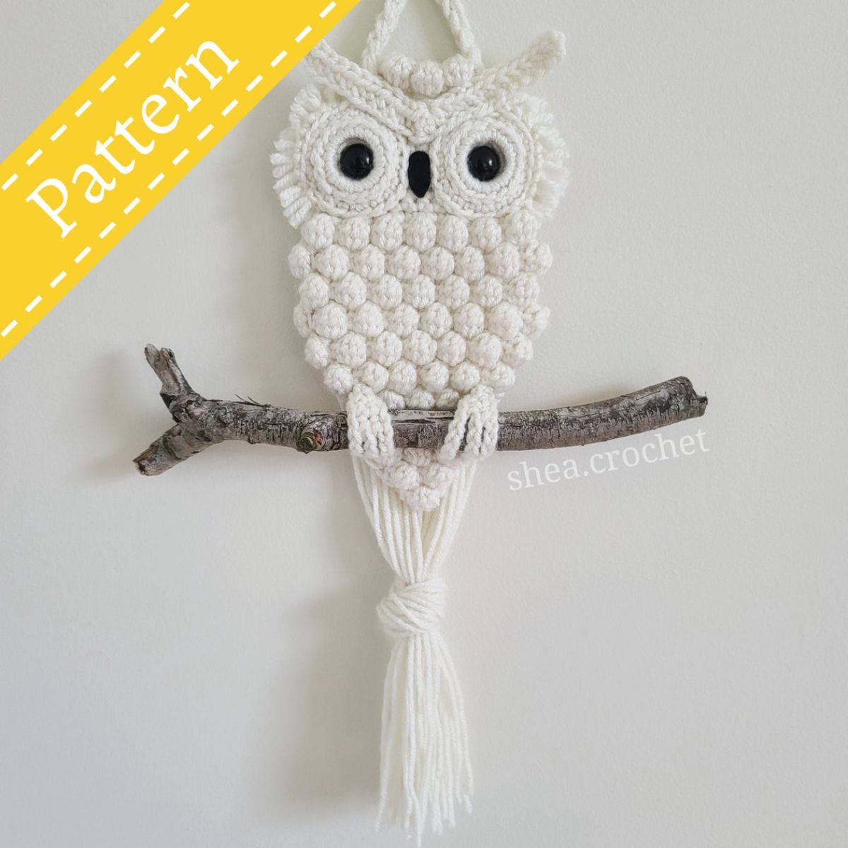 White crochet owl wall hanging with black eyes standing on a branch with long white tassels hanging down from the owl’s feet.