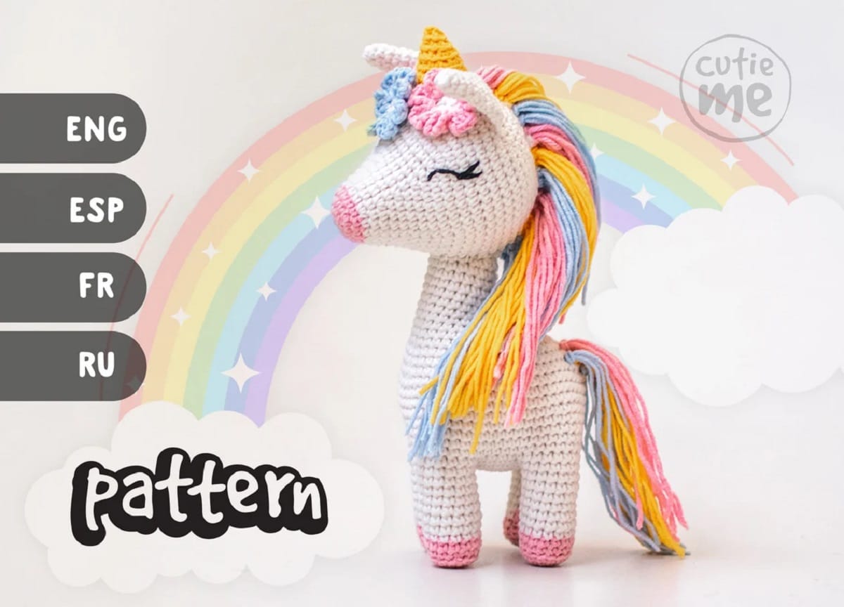 A white crochet stuffed unicorn with a multicolored pastel mane and matching tail with pink and blue flowers around its yellow horn.