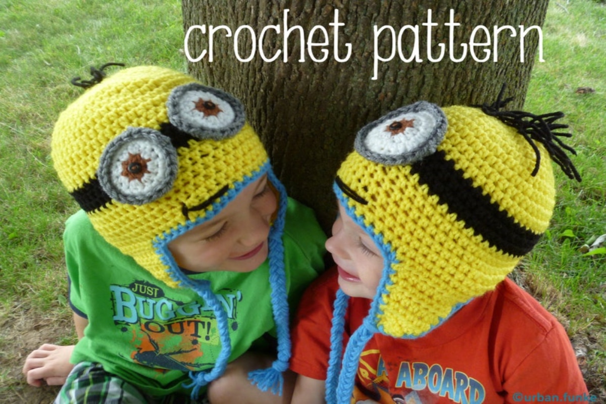  Two young children sat by a tree wearing yellow crochet minion hats, one with two white and gray eyes, the other with one and yellow ear flaps with blue braids hanging from them.