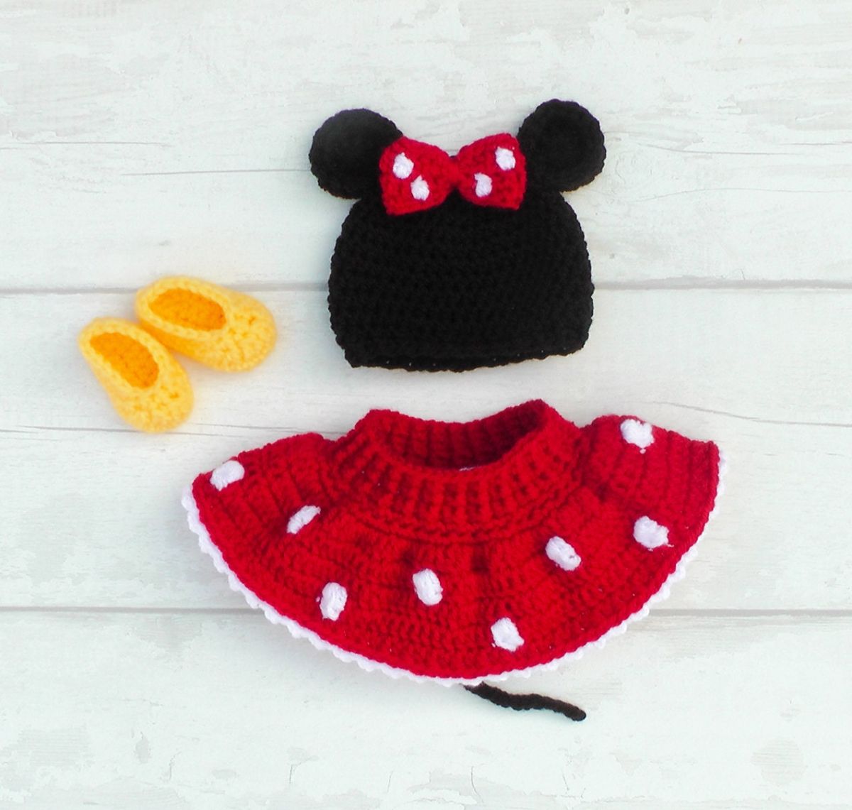 Crochet Minnie Mouse baby costume with a red skirt with white spots, yellow slip on shoes, and a black and white beanie with a red and white bow and mouse ears.