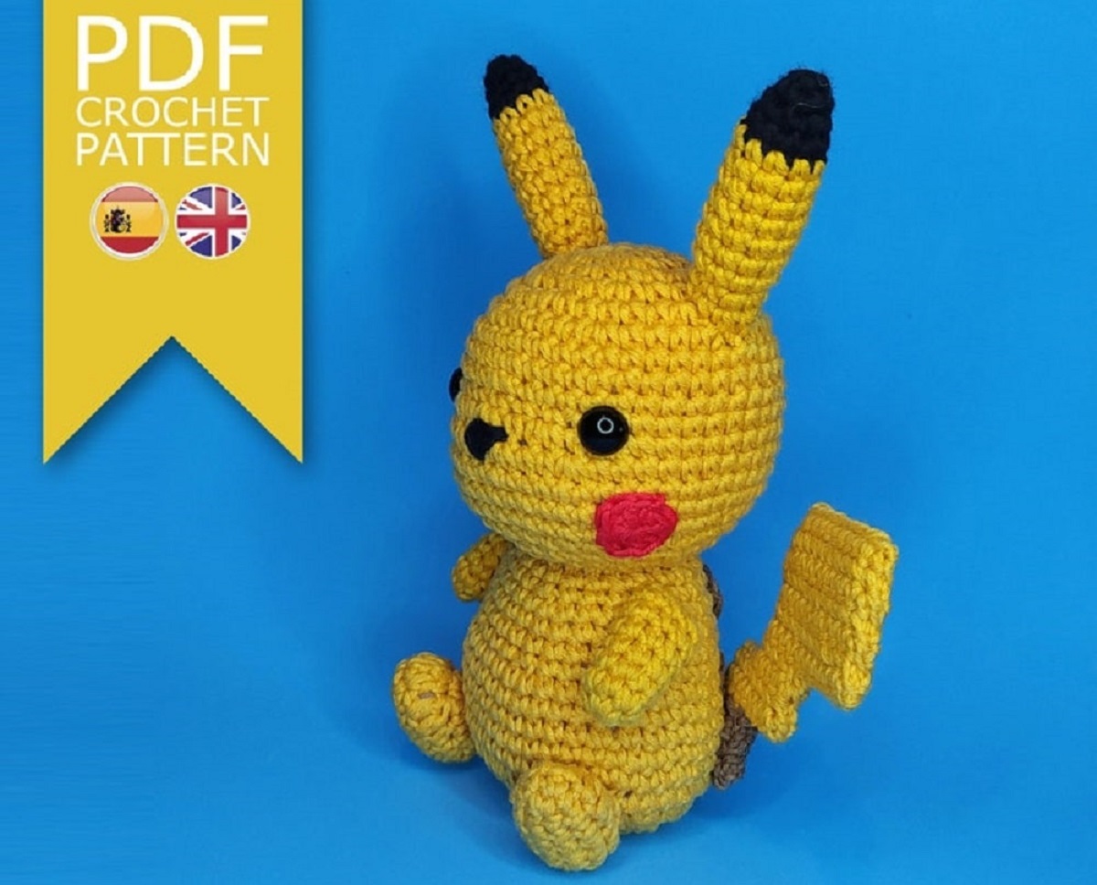 Stuffed crochet Pickachu sitting up on a blue background with black stitching for its eyes and nose and red round cheeks.